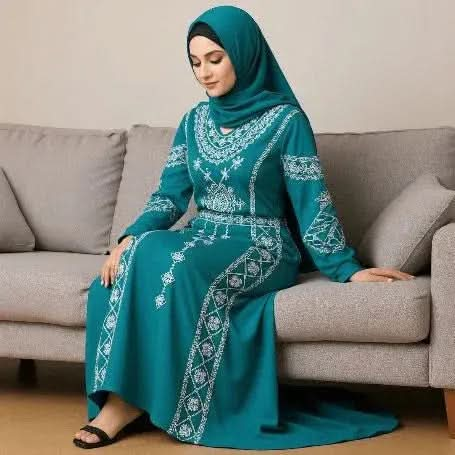 Woman wearing teal abaya dress with white embroidery and matching hijab sitting on gray sofa