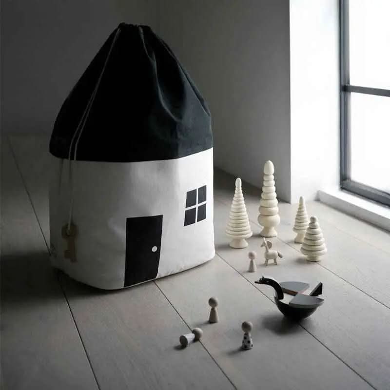 Black and white house-shaped cotton storage bag with wooden toys and trees on light wood floor near window
